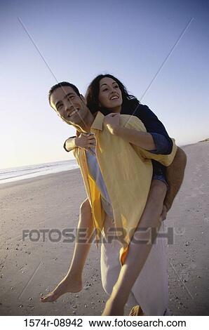 Stock Image - Young woman riding piggyback on a young man at the beach. Fotosearch