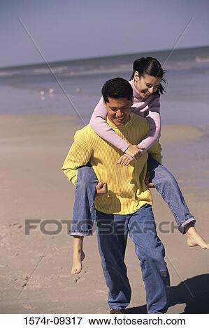 Stock Photo - Young woman riding piggyback on a young man at the beach. Fotosearch