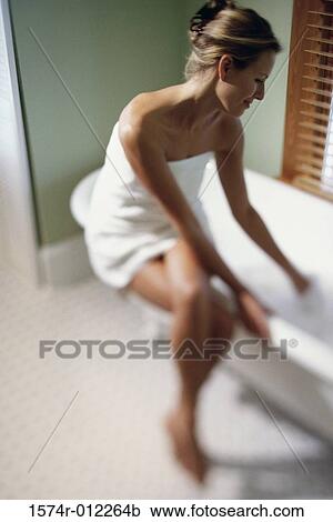 Stock Image - Young woman wrapped in a towel sitting on the edge of a bathtub. Fotosearch