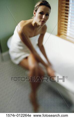 Stock Image - Young woman wrapped in a towel sitting on the edge of a bathtub. Fotosearch