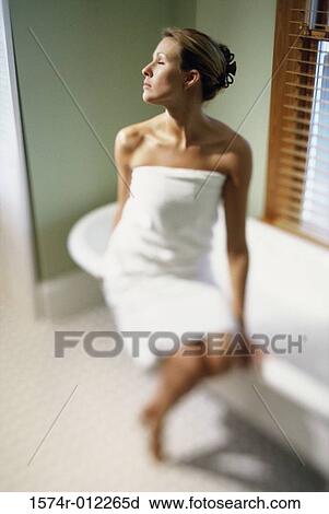 Stock Image - Young woman wrapped in a towel sitting on the edge of a bathtub. Fotosearch