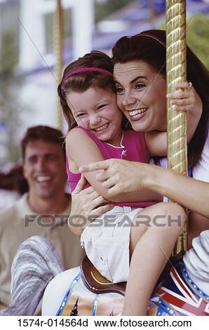 Stock Image - Daughter sitting on a carousel horse with her mother standing beside her. Fotosearch