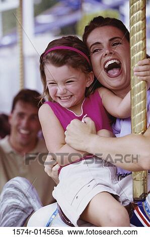 Stock Image - Daughter sitting on a carousel horse with her mother standing beside her. Fotosearch