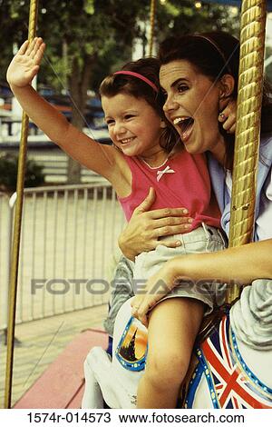 Stock Image - Daughter waving her hand on a carousel horse with her mother standing beside her. Fotosearch