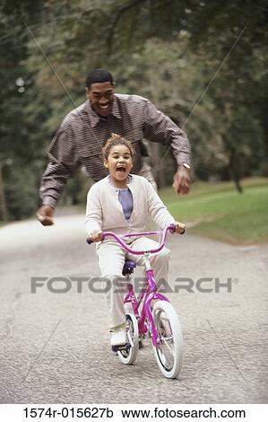 Stock Image - Father helping his daughter ride a bicycle. Fotosearch
