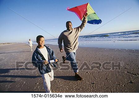 Stock Image - Father running on the beach flying a kite with his son. Fotosearch