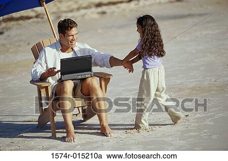 Stock Image - Father working on a laptop with his daughter pulling his hand on the beach. Fotosearch