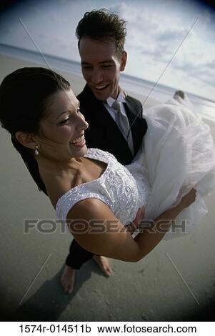 Stock Image - High angle view of a groom carrying his bride on the beach. Fotosearch