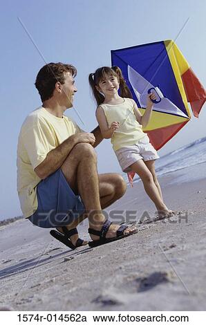 Stock Image - Low angle view of a father helping his daughter fly a kite on the beach. Fotosearch