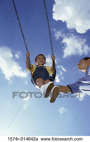 Stock Image - Low angle view of a father pushing his son on a swing. Fotosearch