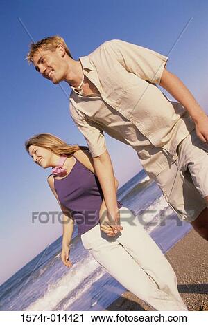Stock Image - Low angle view of a teenage couple holding hands and walking on the beach. Fotosearch