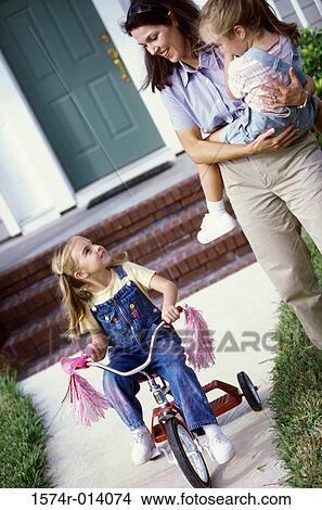 Picture - Mother carrying her daughter looking at her other daughter riding a tricycle. Fotosearch
