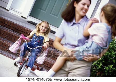 Stock Image - Mother carrying her daughter with another daughter cycling in the background. Fotosearch