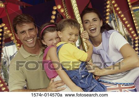 Stock Image - Portrait of a son and daughter sitting on a carousel horse with their parents standing beside them. Fotosearch