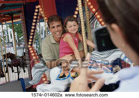 Stock Image - Rear view of a woman taking a photograph of her husband and daughter sitting on a carousel horse. Fotosearch