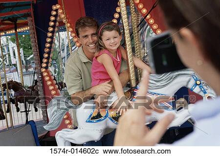 Stock Image - Rear view of a woman taking a photograph of her husband and daughter sitting on a carousel horse. Fotosearch
