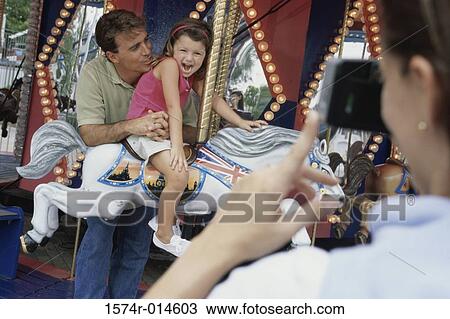 Stock Image - Rear view of a woman taking a photograph of her husband and daughter sitting on a carousel horse. Fotosearch