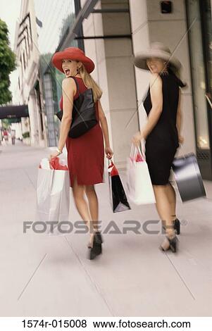 Rear view of two young women carrying shopping bags walking on a sidewalk View Large Photo Image Stock Photo - Rear view of two young women carrying shopping bags walking on a sidewalk. Fotosearch