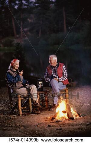 Stock Image - Senior couple sitting near a campfire talking to each other. Fotosearch