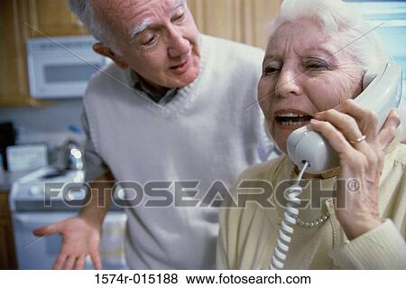 Stock Photo - Senior woman talking on the telephone with a senior man standing beside her. Fotosearch