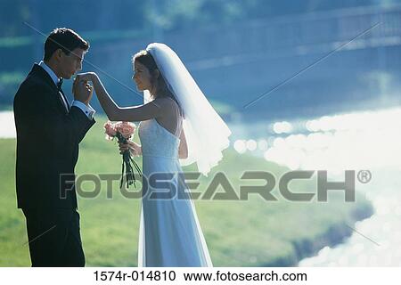 Stock Image - Side profile of a groom kissing his bride's hand near a lake. Fotosearch