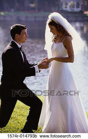 Stock Image - Side profile of a groom proposing to his bride near a lake. Fotosearch