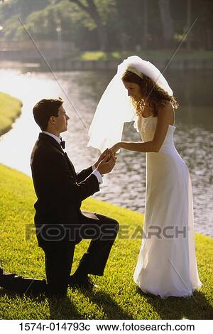 Stock Image - Side profile of a groom proposing to his bride near a lake. Fotosearch