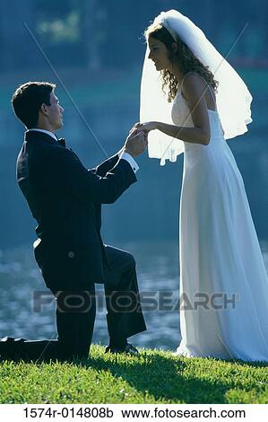 Stock Image - Side profile of a groom proposing to his bride near a lake. Fotosearch