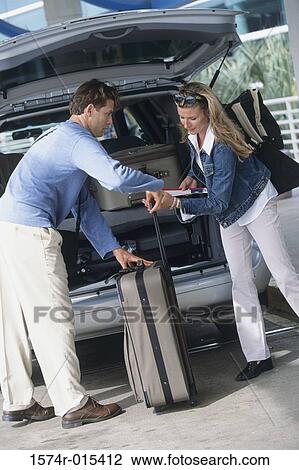 Stock Image - Side profile of a young couple loading luggage into the back of a car. Fotosearch