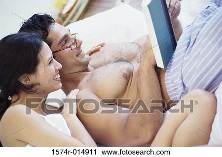 Stock Image - Side profile of a young couple reading a book in bed. Fotosearch