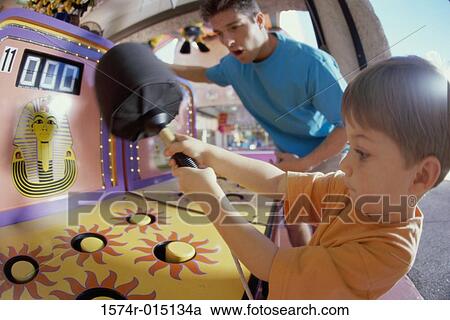 Stock Image - Son playing games with his father standing beside him in an amusement park. Fotosearch