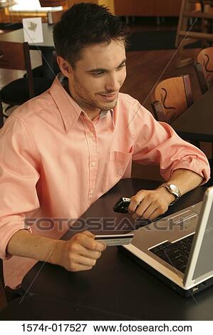 Stock Photo - Young man sitting in front of a laptop holding a credit card. Fotosearch