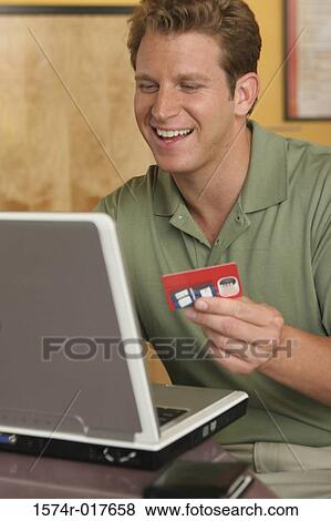 Young man sitting in front of a laptop holding a credit card View Large Photo Image Stock Photo - Young man sitting in front of a laptop holding a credit card. Fotosearch