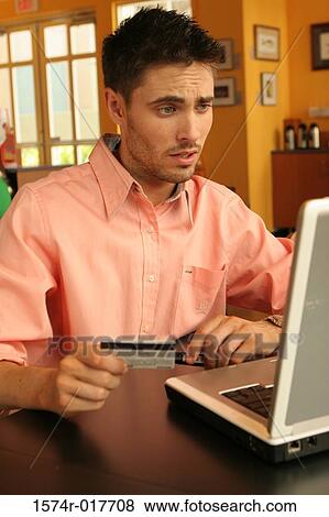 Young man sitting using a laptop holding a credit card View Large Photo Image Stock Photo - Young man sitting using a laptop holding a credit card. Fotosearch