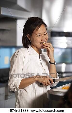 Stock Photo - Close-up of a mid adult woman eating a slice of carrot in the kitchen. Fotosearch