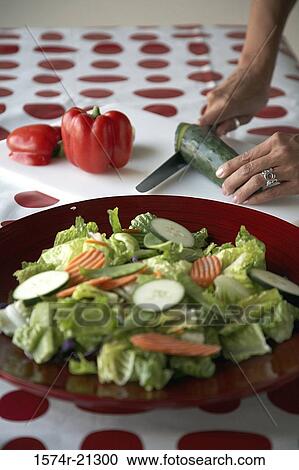 Stock Image - Close-up of a salad on a plate with a person cutting slices of cucumber in the background. Fotosearch