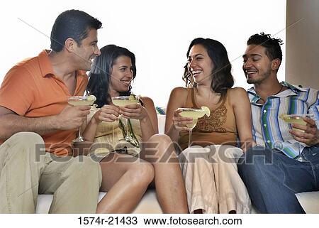 Stock Image - Mid adult couple and a young couple sitting on a couch and holding margaritas. Fotosearch