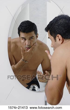 Stock Image - Mid adult man looking at himself in a mirror in the bathroom. Fotosearch