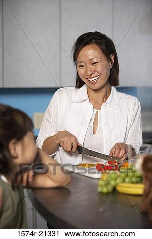 Stock Image - Mid adult woman cutting vegetables on a cutting board looking at her daughter. Fotosearch