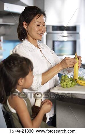 Stock Image - Mid adult woman peeling a banana with her daughter standing beside her in a kitchen. Fotosearch