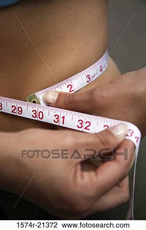Stock Image - Mid section view of a woman measuring her waist with a tape measure. Fotosearch
