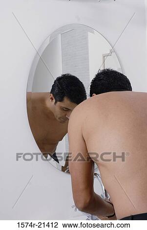 Stock Image - Rear view of a mid adult man washing his face in front of a mirror in the bathroom. Fotosearch