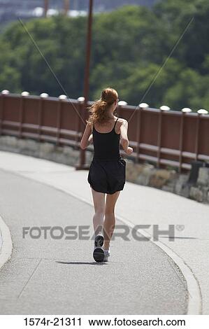 Stock Image - Rear view of a young woman jogging on a road. Fotosearch