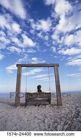 Stock Image - Rear view of a young woman sitting in a swing on the beach. Fotosearch
