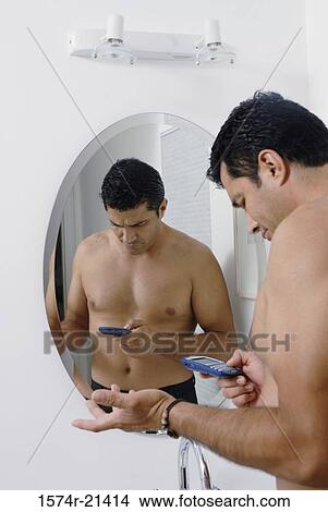 Picture - Side profile of a mid adult man standing in front of a mirror and using a mobile phone in the bathroom. Fotosearch