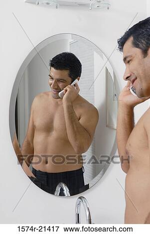 Stock Photo - Side profile of a mid adult man standing in front of a mirror and talking on a mobile phone in the bathroom. Fotosearch