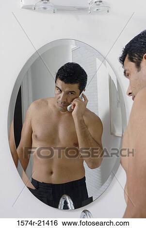 Stock Photograph - Side profile of a mid adult man standing in front of a mirror and talking on a mobile phone in the bathroom. Fotosearch