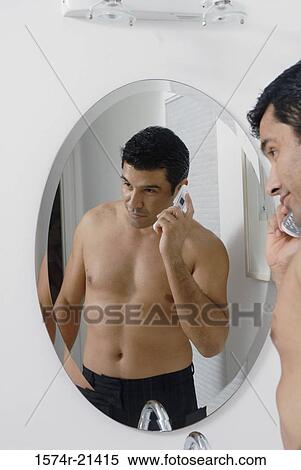 Stock Photography - Side profile of a mid adult man standing in front of a mirror and talking on a mobile phone in the bathroom. Fotosearch