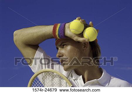 Stock Photo - Young man holding two tennis balls wiping his face with an arm band. Fotosearch