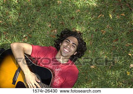 Stock Image - High angle view of a young man lying in grass and playing a guitar. Fotosearch